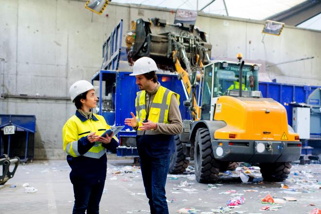 Zwei Arbeiter vor einem Bagger, der einen Abfallcontainer befüllt